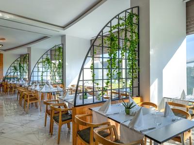 Bright restaurant with wooden tables, chairs, and large windows overlooking plants.