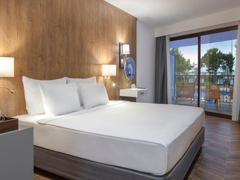 Chambre d'hôtel moderne avec grand lit, mur en bois, lampes de chevet et balcon avec vue sur les arbres.