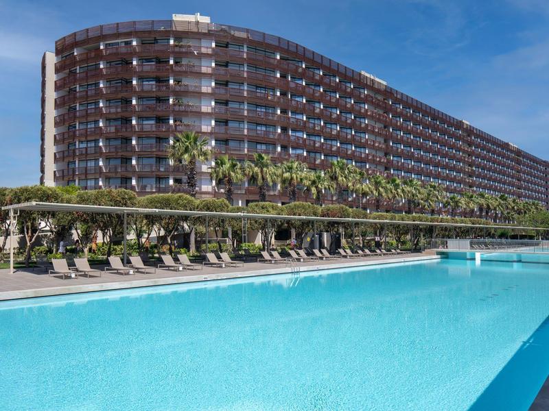 Grand bâtiment hôtelier avec chambres à balcon, piscine et chaises longues sous un ciel ensoleillé