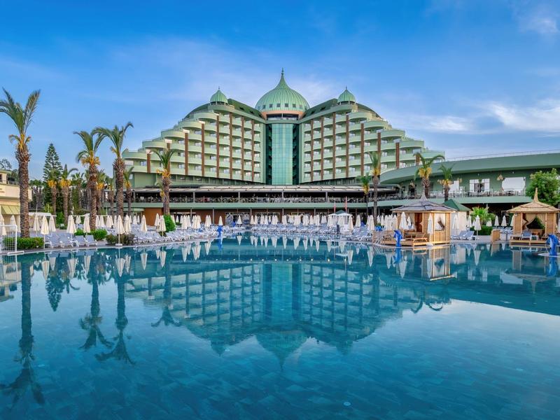 Groot meerlaagshotel met een groot zwembad op de voorgrond onder een blauwe lucht.