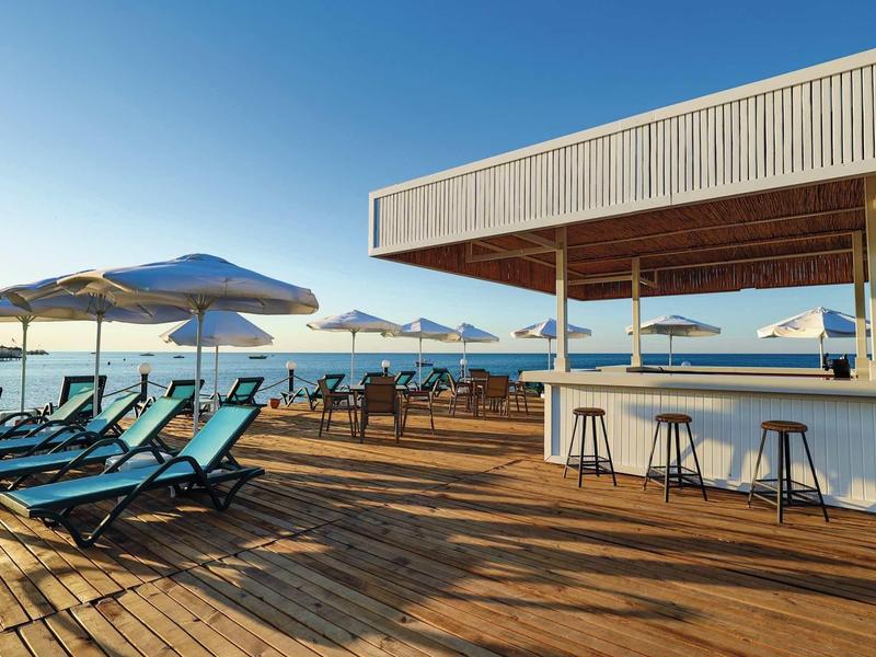 Terraza soleada con tumbonas azules y bar con taburetes junto al mar bajo cielo despejado.