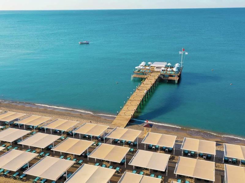 Un muelle largo conduce a una pequeña plataforma en el tranquilo mar azul, frente a filas de sombrillas en la playa.