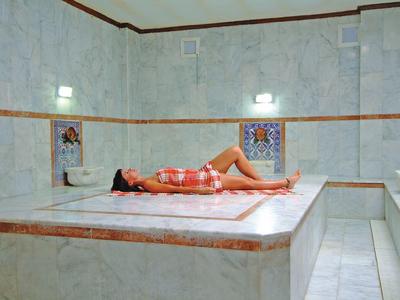 Frau liegt entspannt auf Marmorplatte in hellem, weiß gefliestem Hammam mit Verzierungen.