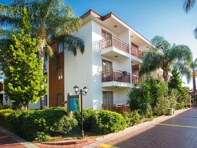 Dreistöckiges Wohnhaus mit Balkonen, umgeben von Palmen und grünem Buschwerk an sonnigem Tag.