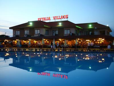 Restaurant mit beleuchtetem Schild "ITALIA VILLAGE" neben großem, ruhigem Pool bei Abenddämmerung.