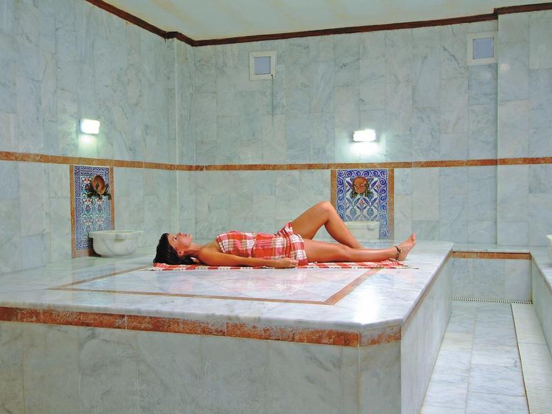 Frau liegt entspannt auf Marmorplatte in hellem, weiß gefliestem Hammam mit Verzierungen.