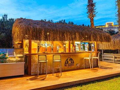 Bar mit Strohdach, Holztheke und mehreren Stühlen bei Dämmerung im Freien.