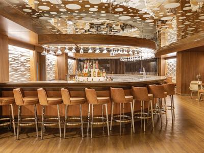 Moderne Bar mit hölzernen Stühlen, rundem Bartresen, Spiegeln und dekorativer Decke mit Fleckenmuster.