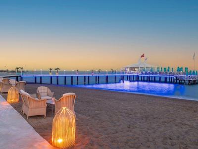 Strand mit Sand, Korbstühlen, Laternen und einem beleuchteten Pier bei Sonnenuntergang.