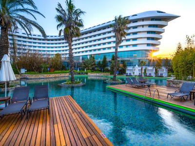 Moderní hotelový komplex se bazénem, dřevěnými terasami, palmami a západem slunce v pozadí.