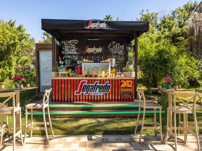 Kleiner, offener Cafékiosk mit hohen Stühlen, Pflanzen und sonnigem Himmel im Hintergrund.