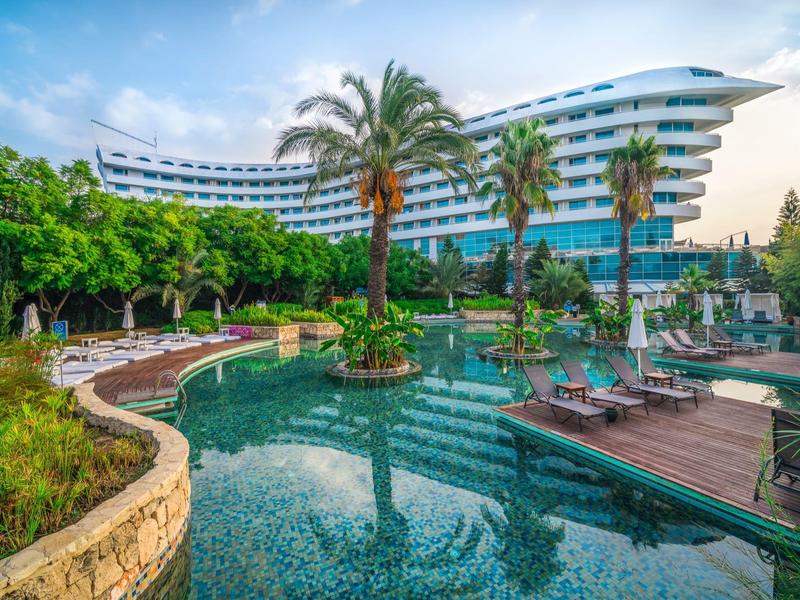 Moderner gekrümmter Hotelbau mit großem Pool, Palmen und Sonnenterrasse unter blauem Himmel.