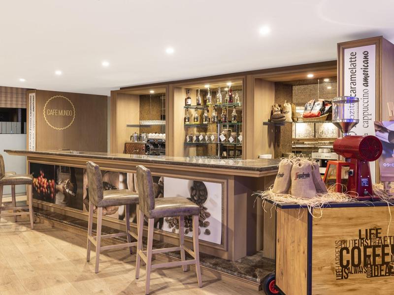 Gemütliche Bar mit Holztheke, Barhockern, Regalen voller Flaschen und einem Kaffeewagen rechts.