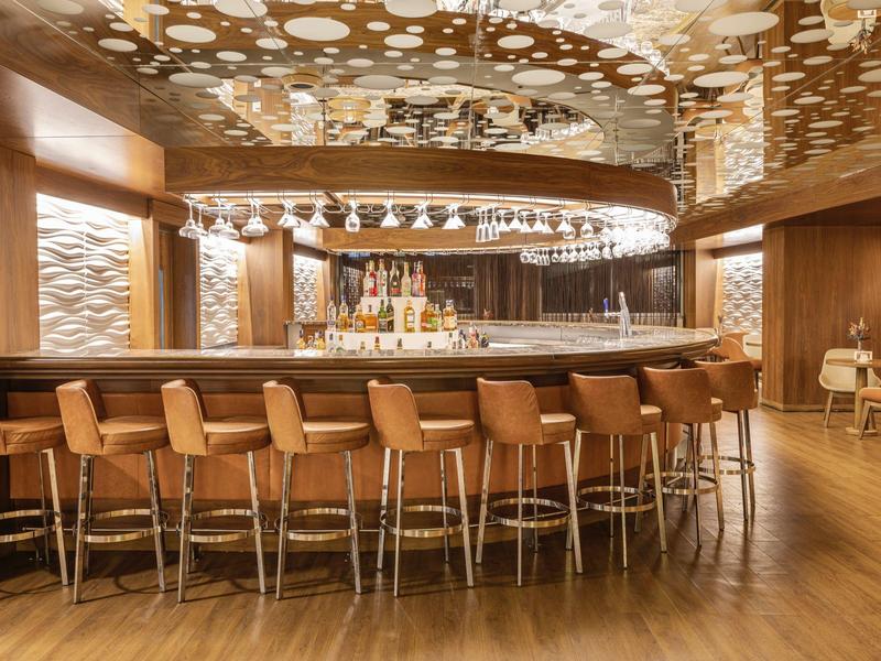 Moderne Bar mit hölzernen Stühlen, rundem Bartresen, Spiegeln und dekorativer Decke mit Fleckenmuster.