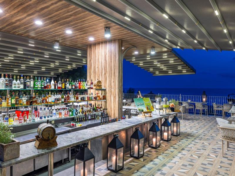 Moderner, offener Barbereich mit Holzdecke, beleuchtetem Tresen und Meerblick bei Abenddämmerung.