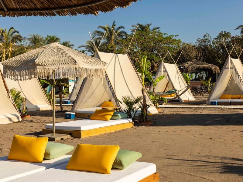 Strand mit weißen Zelten, Liegen mit gelben Kissen und Palmen unter blauem Himmel.