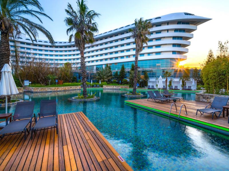 Modernes Hotel mit geschwungenem Gebäude, Pool, Holzterrassen und Palmen bei Sonnenuntergang.