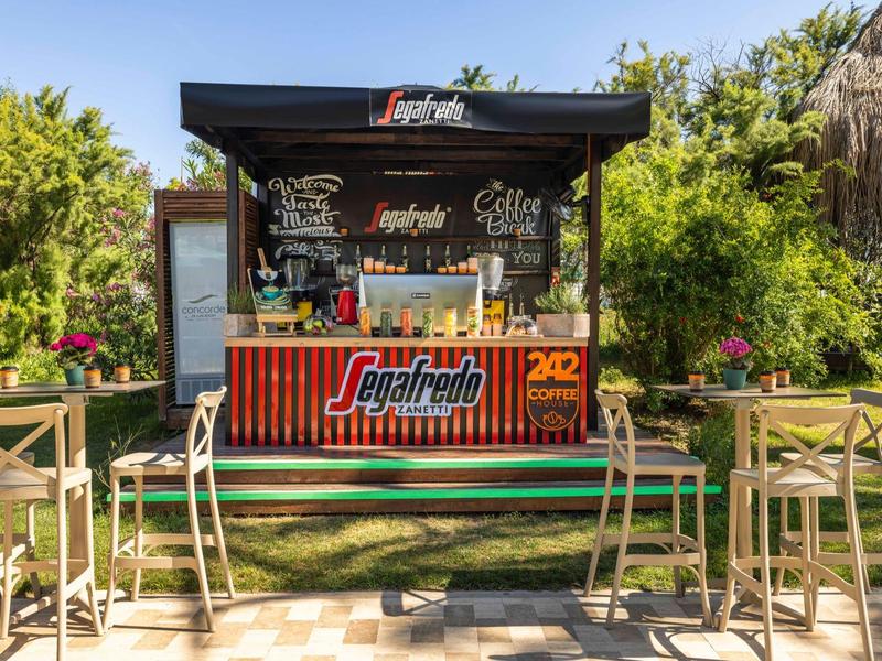 Kleiner, offener Cafékiosk mit hohen Stühlen, Pflanzen und sonnigem Himmel im Hintergrund.
