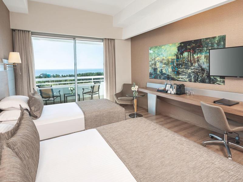 Modernes Hotelzimmer mit zwei Betten, Balkon, Meerblick und Arbeitsplatz mit Flachbildschirm.