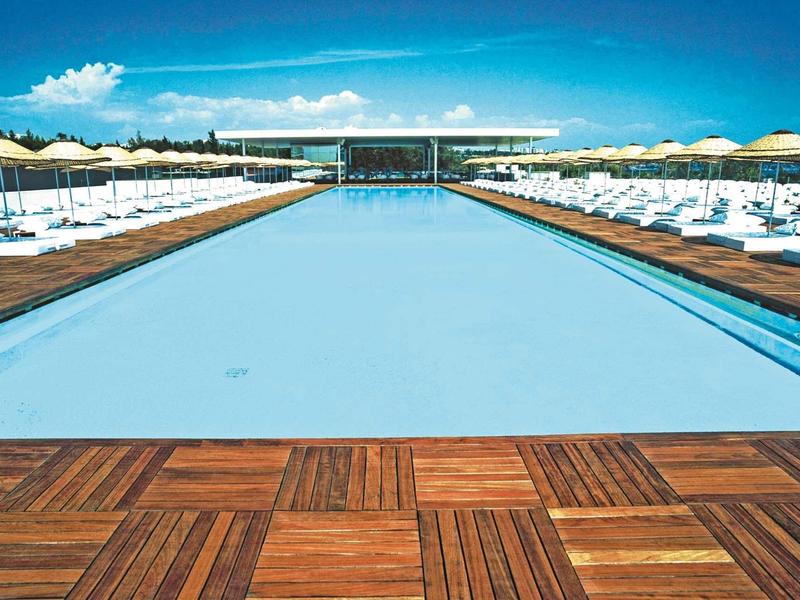 Großer Pool mit hellblauem Wasser, weißen Liegestühlen und Sonnenschirmen auf Holzboden.