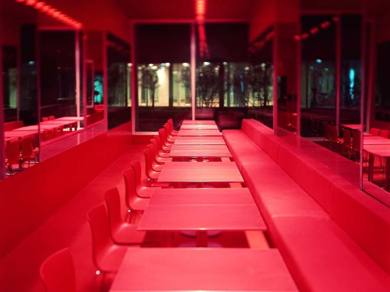 Leerer, langer Raum mit roten Wänden, Tischen, Stühlen und beleuchteter Decke.