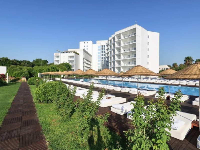 Moderner, weißer Hotelkomplex mit Pool, Liegen und Sonnenschirmen unter blauem Himmel.
