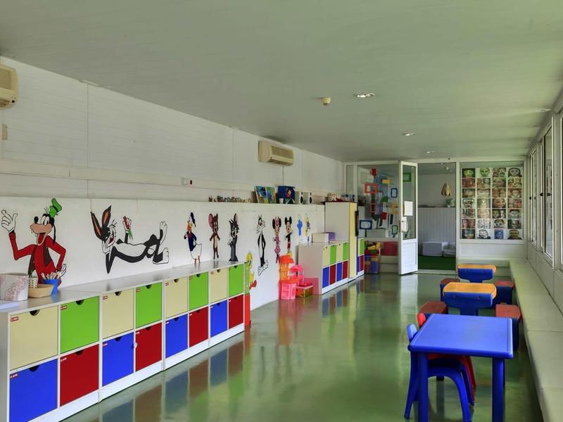 Hell eingerichteter Spielraum mit bunten Regalen, Kindertischen und Fenstern an der Seite.