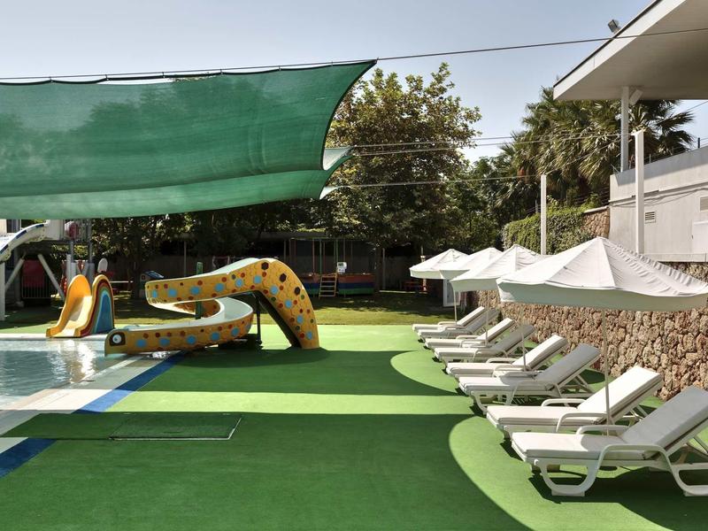 Spielplatz mit Wasserrutsche, Sonnenschirmen und Liegestühlen auf grünem Kunstrasen.