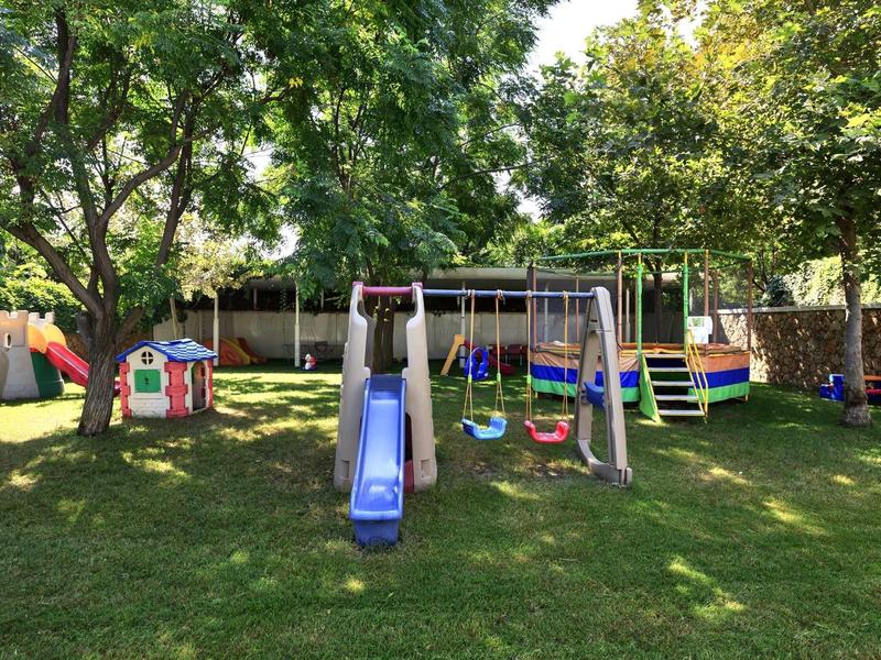 Spielplatz mit Rutsche, Schaukeln, Klettergerüst und Spielhäuschen im Grünen unter großen Bäumen.