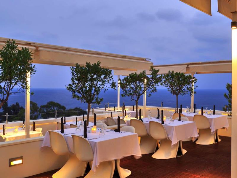 Terrassenrestaurant mit weißen Tischen, Stühlen und kleinen Bäumen bei Abenddämmerung am Meer.