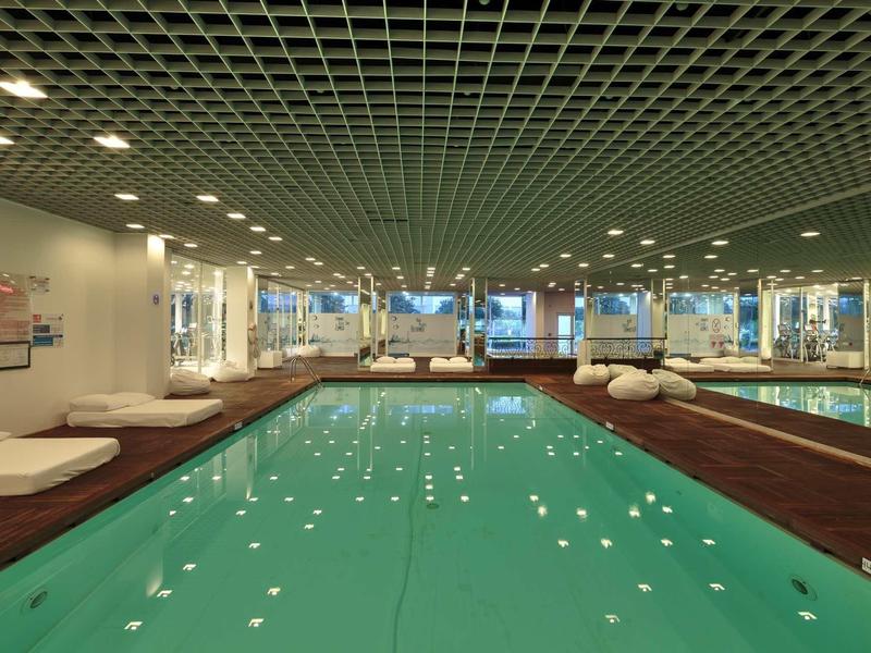 Innenraum mit großem Schwimmbecken, Holzdeck, weißen Liegen und Decke mit quadratischem Raster.