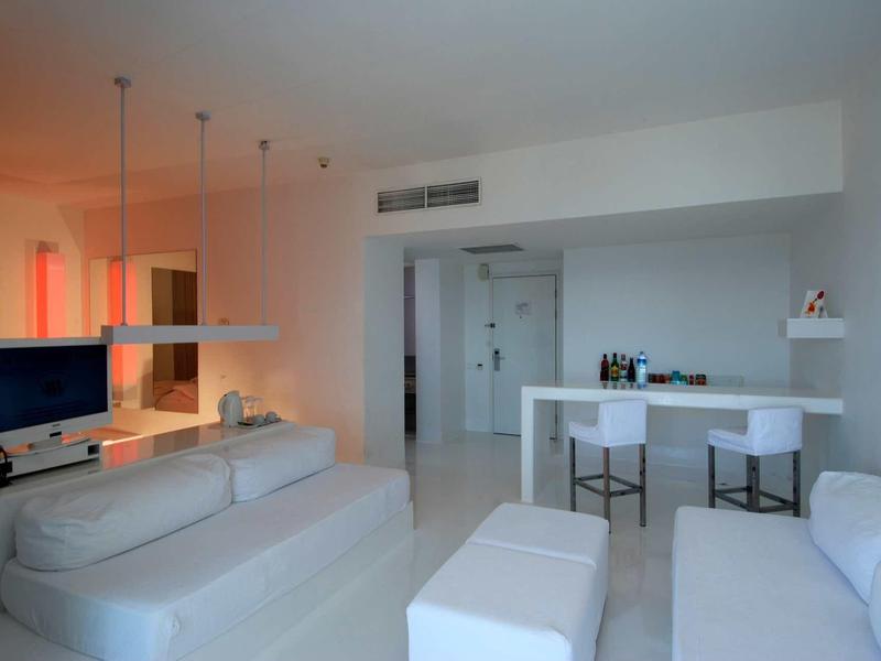 Helles Wohnzimmer mit weißen Möbeln, Sofa, Barhockern, TV und moderner Beleuchtung.