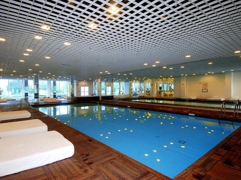 Innenbereich mit rechteckigem Schwimmbecken, Holzfußboden, weißen Liegen und Spiegelwand.