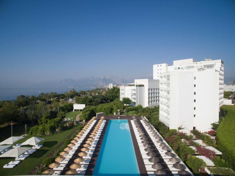 Großer rechteckiger Pool mit Liegestühlen und Sonnenschirmen neben weißen Hochhäusern und grünem Park.