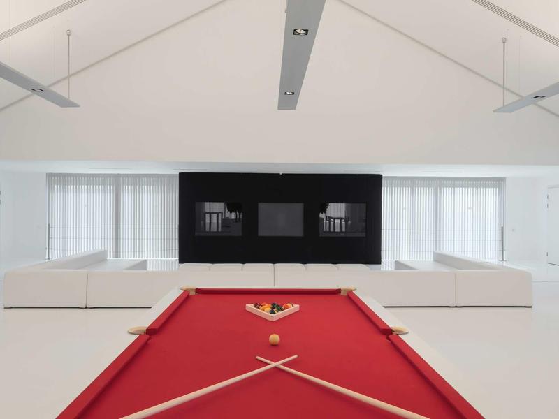 Moderner Raum mit rotem Billardtisch, weißen Sofas, großen Fenstern und hellen Wänden und Decke.