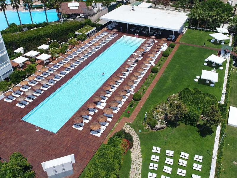 Großer Außenpool mit blauen Wasser, umgeben von weißen Liegestühlen und grünem Rasen.