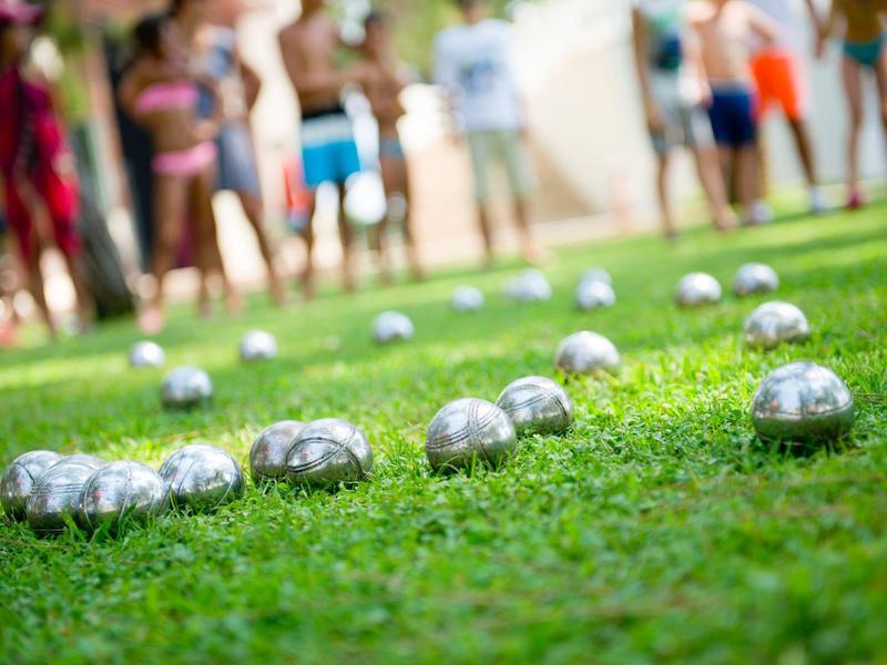 Metallkugeln auf grünem Rasen, unscharfe Menschen im Hintergrund, Spiel bei sonnigem Wetter.