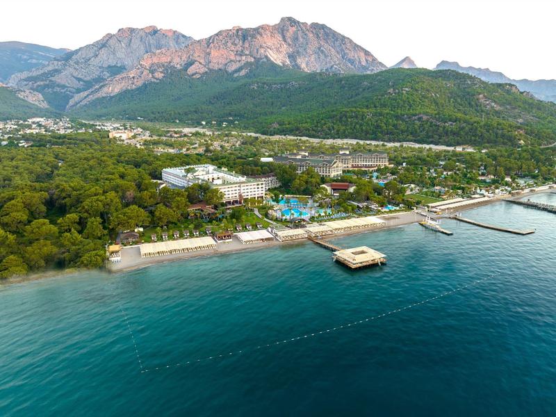 Bucht mit blauem Meer, Strand, grünen Bäumen und Bergen im Hintergrund bei klarem Himmel.