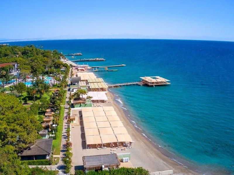 Küste mit Sandstrand, grünen Bäumen und mehreren Liegestuhlreihen am blauen Meer unter klarem Himmel.