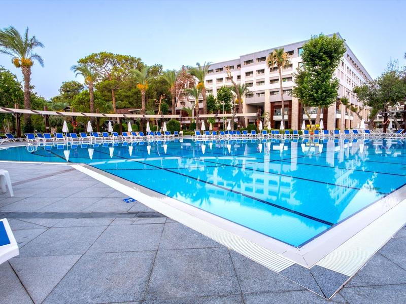 Großer blauer Pool mit Liegestühlen und Sonnenschirmen vor einem mehrstöckigen Hotelgebäude.