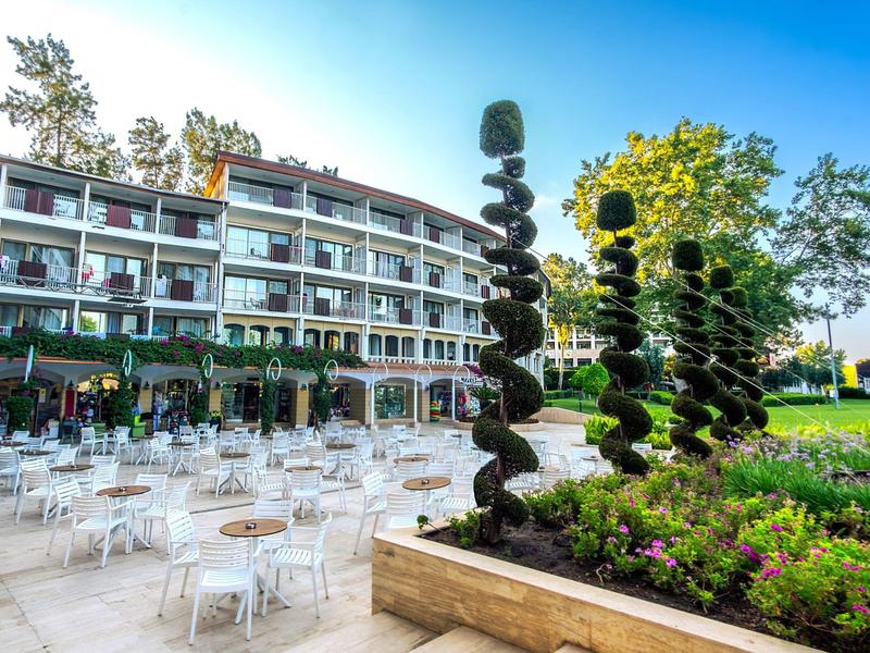 Großes Hotel mit Balkonzimmern, davor viele weiße Tische und Stühle, rechts Spiral-Skulpturen im Garten.