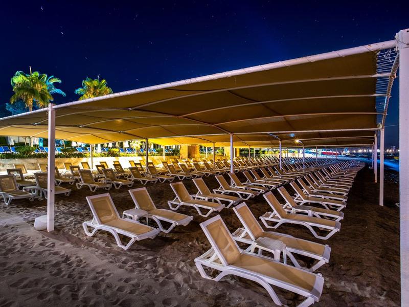 Viele leere Liegestühle unter einem großen Sonnensegel am Strand bei Nacht.