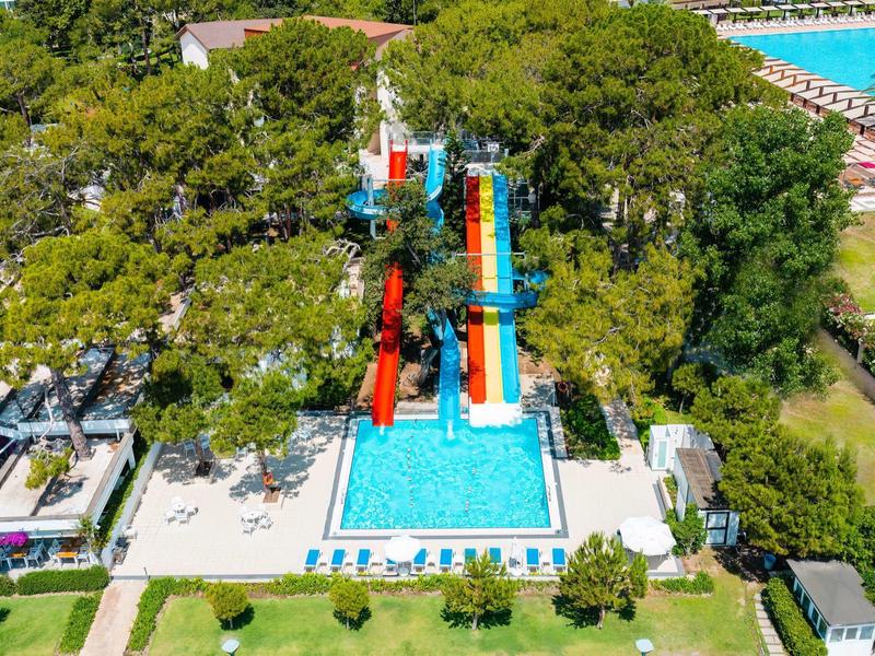 Freibad mit mehreren bunten Wasserrutschen und blauem Becken, umgeben von grünen Bäumen und Liegestühlen.