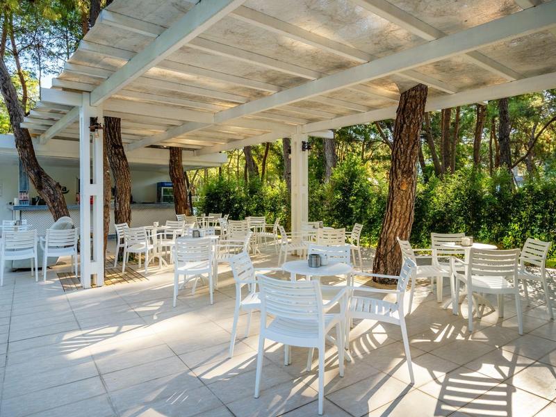 Freiluft-Café mit weißem Holzpergola, weißen Stühlen und Tischen umgeben von Bäumen.