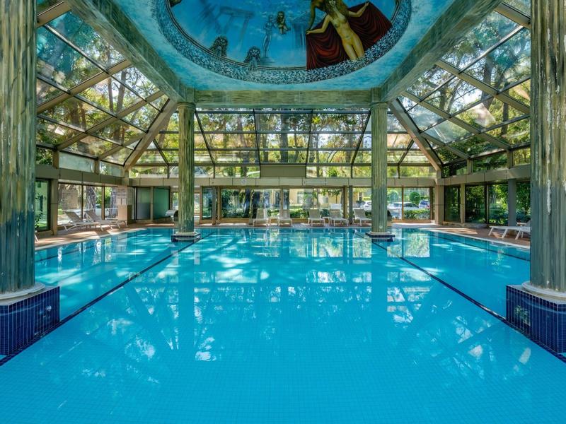 Hallenbad mit blauem Wasser, Glasdach, großen Säulen und Spiegelungen im Wasser.