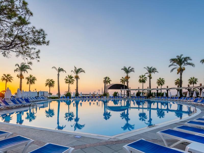 Großer Pool mit blauer Wasseroberfläche, umgeben von Palmen und blauen Liegestühlen bei Sonnenuntergang.
