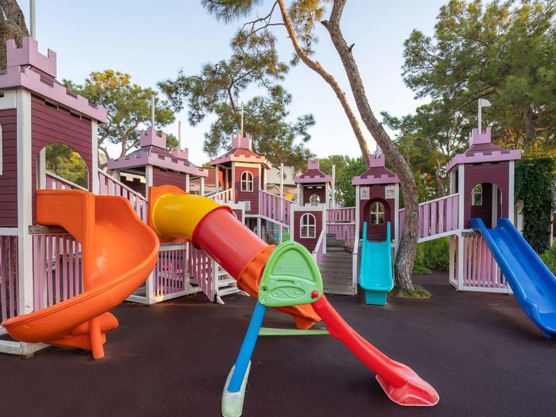 Bunter Spielplatz mit mehreren Rutschen und kleinen Türmen in lila und orange unter Bäumen.
