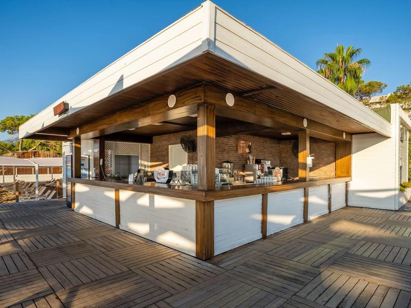 Modernes, offenes Strandcafé mit Holzverkleidung, großem Thekenbereich und Sonnenschein am blauen Himmel.