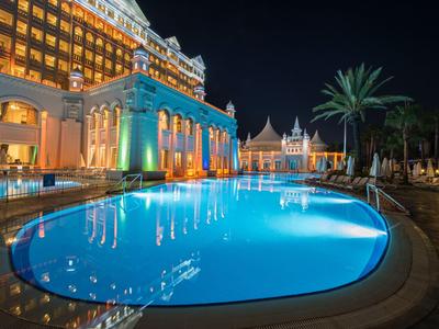 Beleuchteter Hotelpool bei Nacht mit Palmen und reflektierendem Wasser.