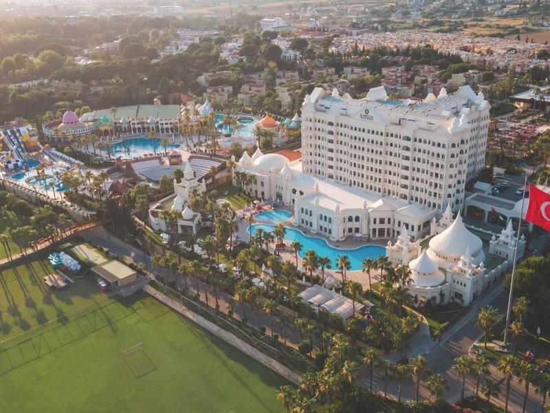 Widok z lotu ptaka na duży ośrodek z kilkoma basenami i białym budynkiem hotelowym obok boiska do piłki nożnej.
